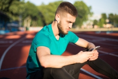 Portrait of man in wireless earphones thoughtfully using cellphone while spending time on running track of stadium